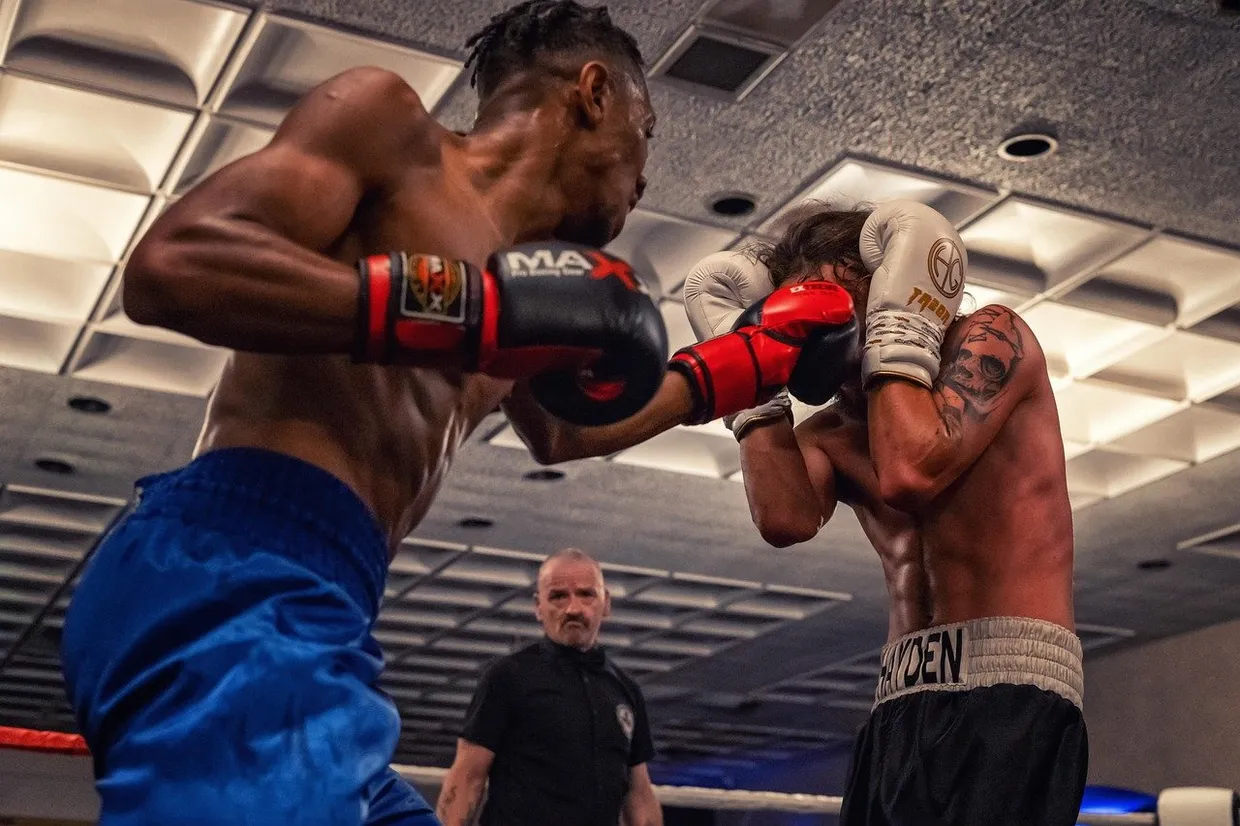 Un boxeador lanza un golpe a la cabeza mientras el otro se cubre con la guardia y el árbitro los observa desde el fondo del ring