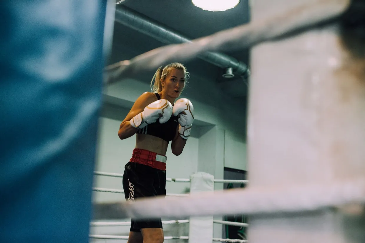 Boxeadora en guardia dentro de un ring de boxeo, con guantes blancos, vista entre las cuerdas