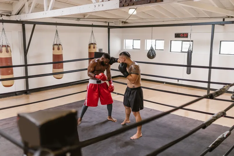 Dos boxeadores entrenando en el ring de un gimnasio, uno cubriéndose la cara y el otro golpeando a corta distancia