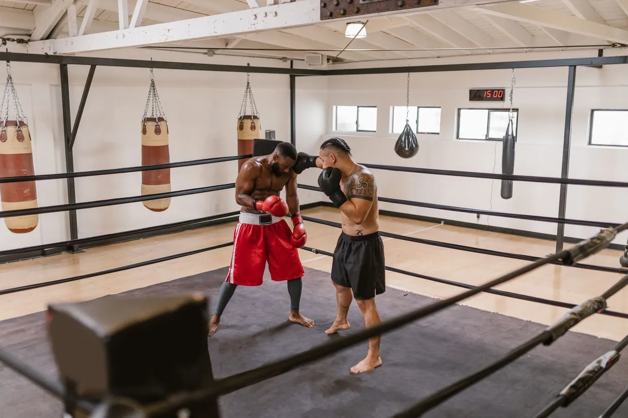 Dos boxeadores entrenando en el ring de un gimnasio, uno cubriéndose la cara y el otro golpeando a corta distancia
