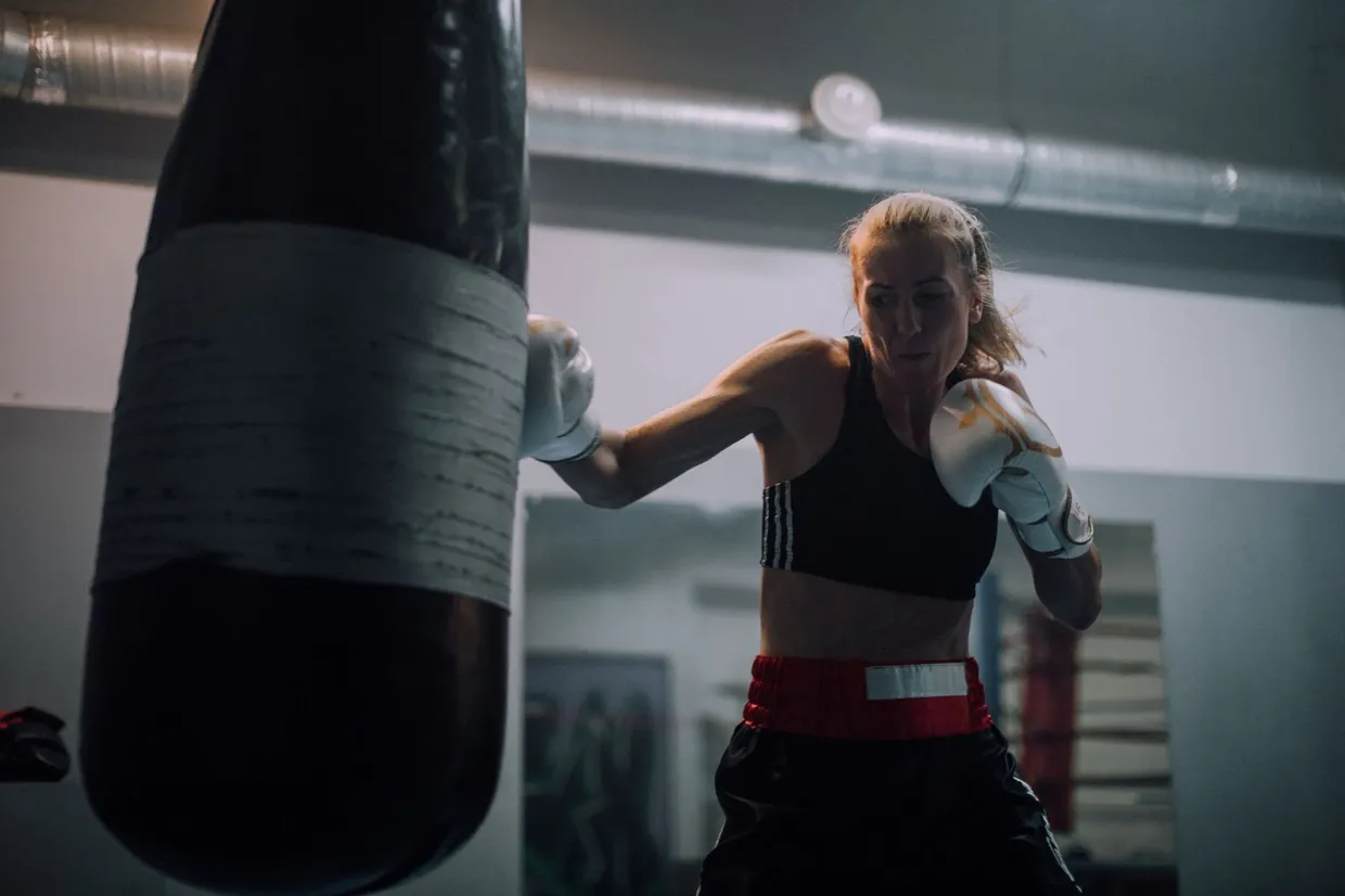 Boxeadora con guantes blancos golpeando un saco pesado en un gimnasio, con la mano de atrás recogida en guardia