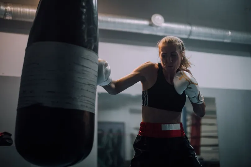 Boxeadora con guantes blancos golpeando un saco pesado en un gimnasio, con la mano de atrás recogida en guardia
