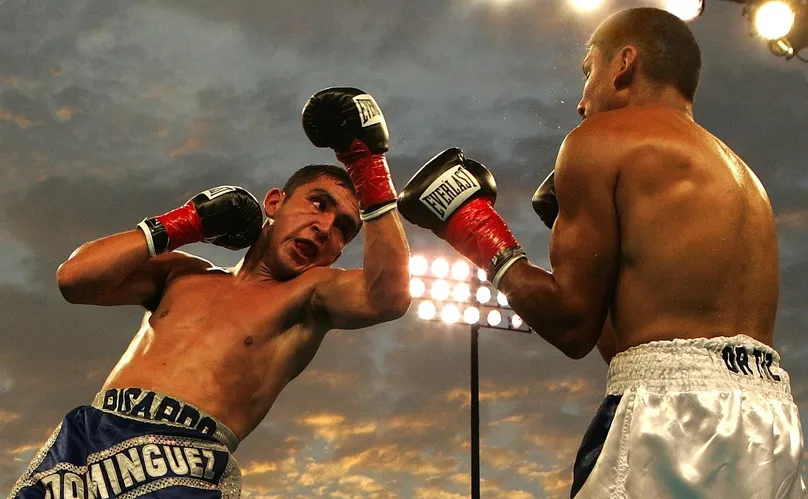 Dos boxeadores profesionales intercambiando golpes en un ring al aire libre, de noche y bajo los focos