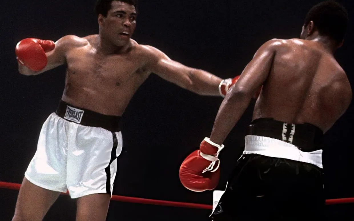 Muhammad Ali, con pantalón blanco, estira el brazo izquierdo hacia la cara de su rival en el ring
