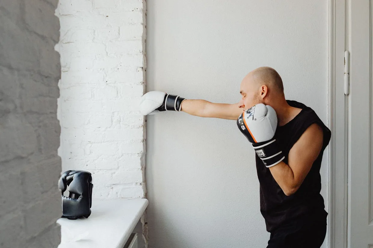 Hombre entrenando boxeo en casa, lanzando un golpe recto frente a una pared blanca con la mano de atrás en la barbilla