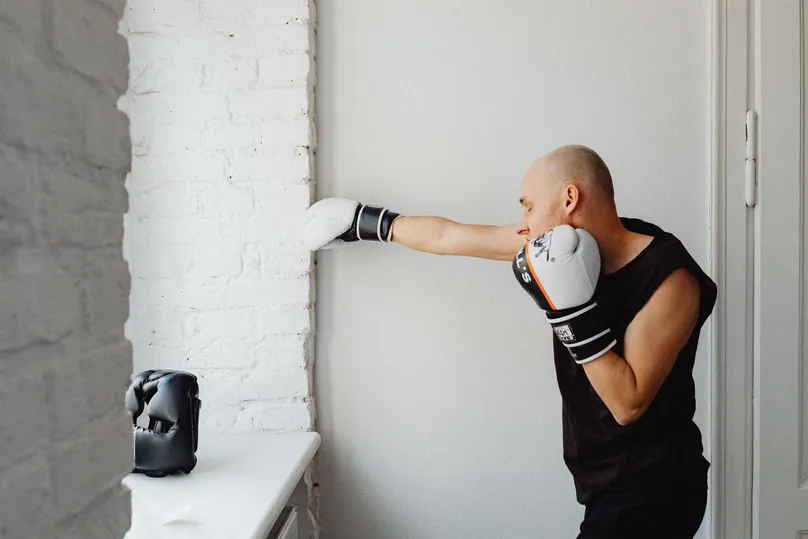 Hombre entrenando boxeo en casa, lanzando un golpe recto frente a una pared blanca con la mano de atrás en la barbilla